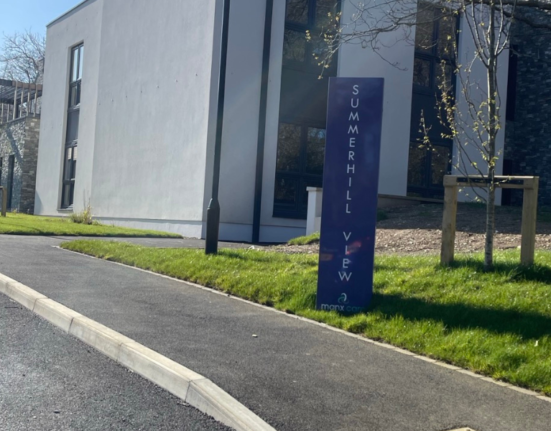 Summerhill View, a care home on the Isle of Man, picture shows the entrance driveway with black tarmac, the green garden and welcome sign.