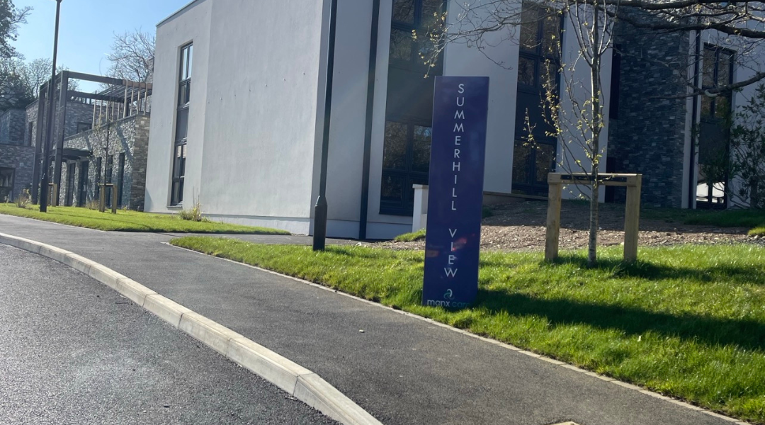 Summerhill View, a care home on the Isle of Man, picture shows the entrance driveway with black tarmac, the green garden and welcome sign.